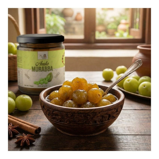 A jar of AMLA Murabba  sits on a rustic wooden table next to a carved brown bowl filled with glossy, golden-yellow mango chutney and a silver spoon. Fresh green mangoes, cinnamon sticks, and star anise are arranged around the table, with a soft-focus window and garden background.