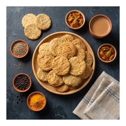 A top-down styled food scene showing a large wooden plate piled with round mathri, surrounded by small clay bowls of spices, seeds, and a cup of tea on a dark textured surface.