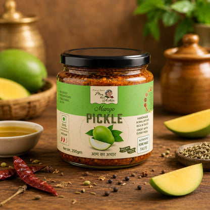 A jar of Mango Pickle by Maa Kaa Achar is displayed on a rustic wooden table surrounded by ingredients like sliced mango, dried red chilies, peppercorns, and spices.