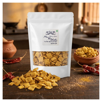 A white stand-up pouch of Maa Made of Thikee Papdi displayed on a rustic wooden table with scattered crackers, red chili peppers, and a small bowl of crackers in the background.