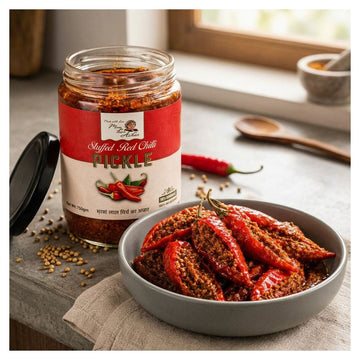 A jar of Pickle Stuffed Red Chili next to a gray bowl filled with red chili peppers in a spicy sauce, set on a rustic stone surface with scattered seeds and a wooden spoon in the background.