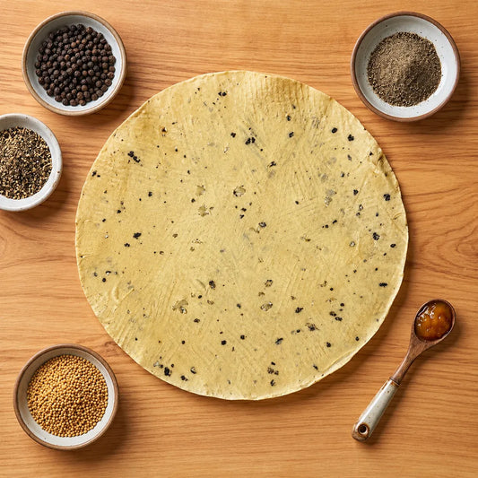 A round handmade moong papad spread on a wooden surface, surrounded by small bowls of spices or seeds and a spoon with a brown spread, arranged in a styled overhead composition.